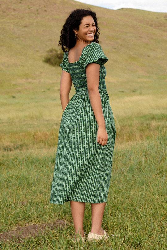 A woman in Ivy City Co’s Sunday Best Dress in Pine Felicity Floral stands smiling over her shoulder in a grassy field. Her curly hair frames her face and she wears light-colored shoes. The nursing and maternity-friendly design of the dress ensures comfort as she enjoys the peaceful landscape, with a gentle hill and clear sky behind her.