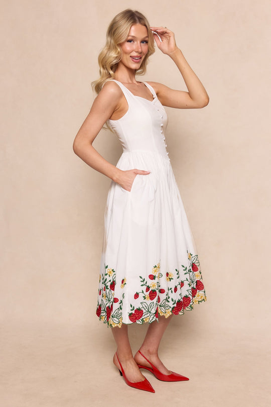 A woman stands smiling against a beige background, one hand touching her hair, wearing the Ivy City Co Clarissa Dress in Strawberry—a white sleeveless dress featuring colorful embroidered floral details at the hem—paired with red pointed-toe heels.
