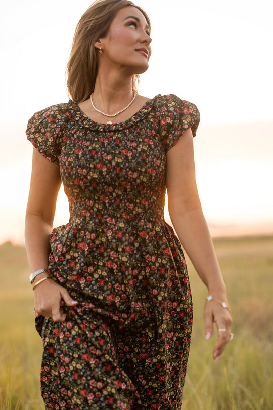 A woman wearing the Annalise Dress in Obsidian Vintage Poppies by Ivy City Co stands outdoors in a grassy field at sunset, gazing off to the side as the light illuminates her face and hair. This maternity-friendly style adds a romantic touch to the serene scene.