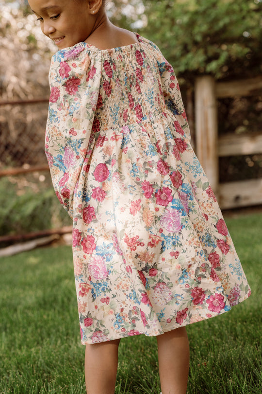 A young child stands outside on the grass, looking down and smiling, wearing the Ivy City Co Mini Adelpha Dress in Autumn Amity Floral. The dress features a smocked bodice and balloon sleeves adorned with a charming floral pattern. A wooden fence and lush greenery create a picturesque background.