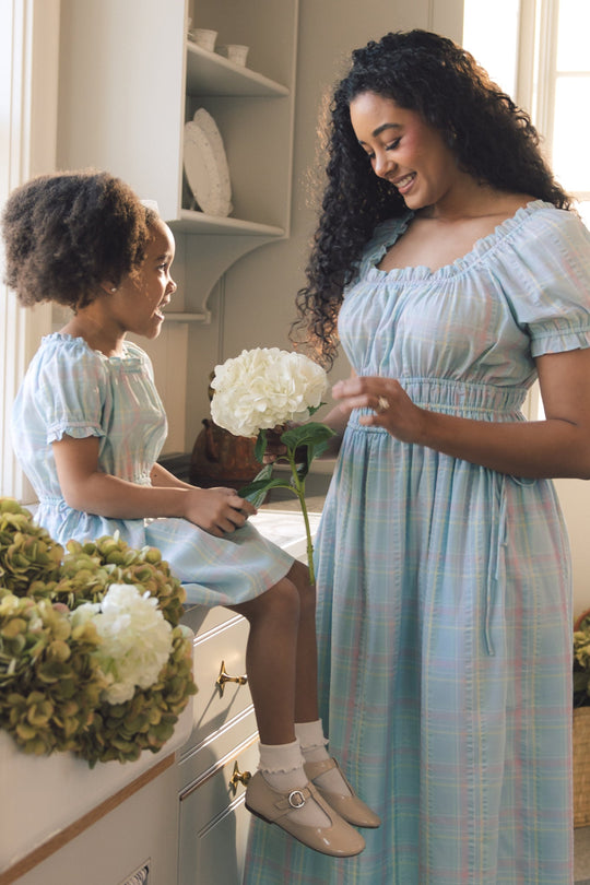 A woman and a young girl, both wearing the Ivy City Co Thalia Dress in Pastel Plaid, smile at each other in a sunlit kitchen while holding white hydrangea flowers.