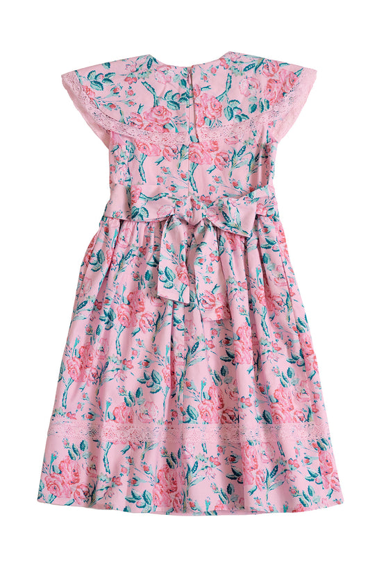 The Mini Aster Dress in Pink Sketched Flower by Ivy City Co features a light pink background adorned with green and pink sketched floral patterns, delicate lace trim, short flutter sleeves, and a charming bow tied at the back—making it an adorable girls’ dress for any special occasion.