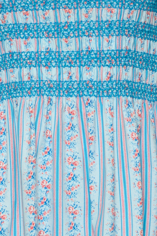A close-up of the Ivy City Co Sunday Best Dress in Rosette Linear Stripe showcases its light blue cotton midi design with vertical red and white stripes and delicate pink floral details, finished with blue floral smocking at the top—thoughtfully crafted to be maternity friendly for easy, effortless summer style.