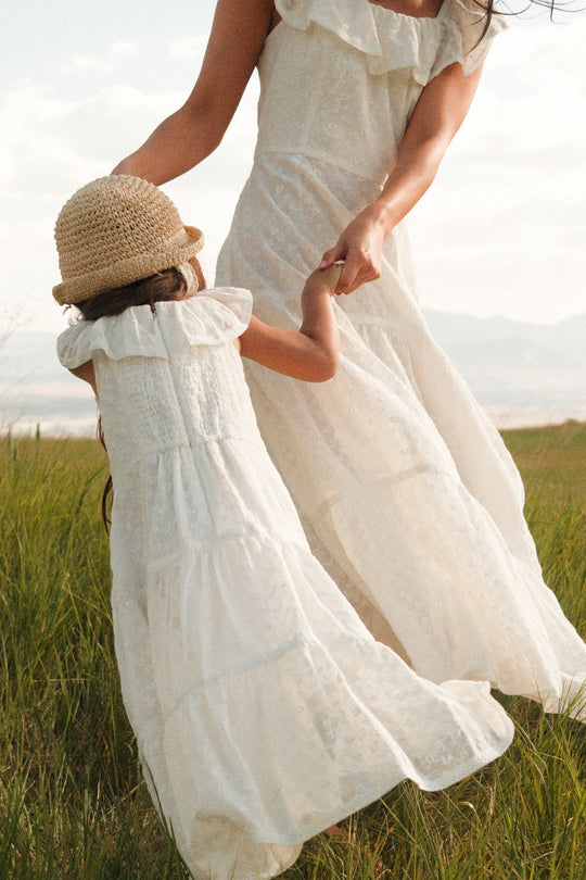 A woman and a young girl twirl hand-in-hand through a sunlit field, both wearing the Alaina Dress in Ivory by Ivy City Co. The smocked bodices of their matching dresses add charming detail, and the girl’s outfit is styled maxi length with a straw hat, capturing the warmth and joy of the outdoors.