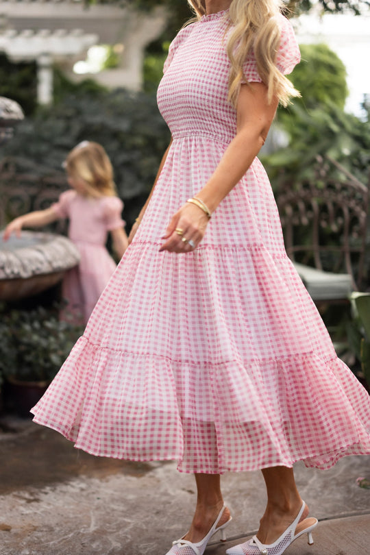 A woman strolls outdoors in the Ivy City Co Cordelia Dress in Desert Rose Gingham, her blonde hair flowing. In the background, a young girl wears a matching dress as she stands by a fountain framed with greenery. The look exudes effortless charm and is maternity friendly.