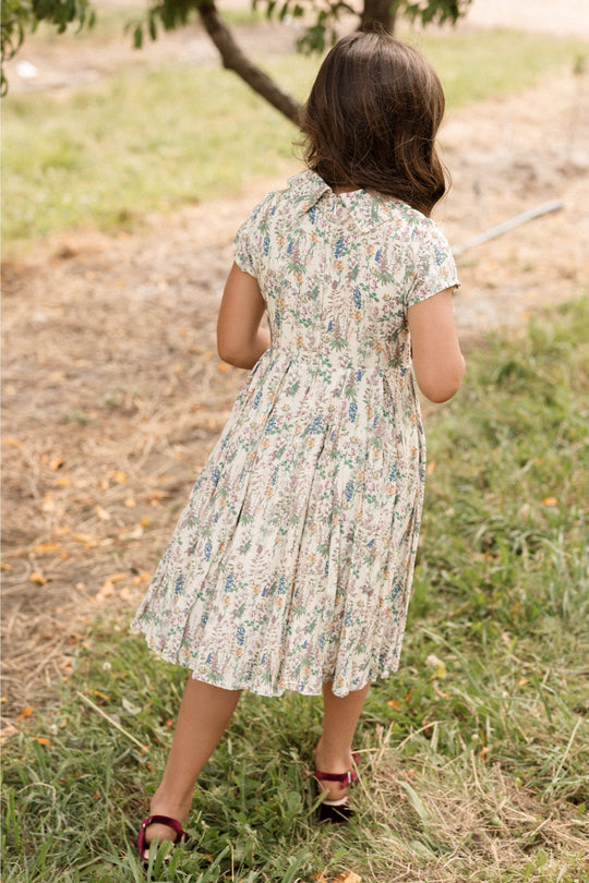 A young girl wearing the Ivy City Co Mini Amber Dress in Autumn Bouquet and sandals walks on grass and dirt, facing away from the camera toward a tree on a sunny day.