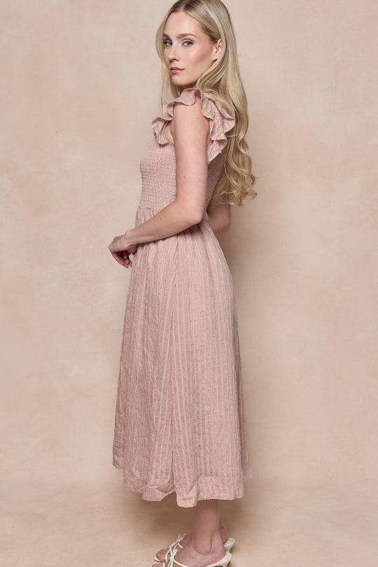 A woman with long blonde hair stands sideways against a beige background, wearing the Ivy City Co Hattie Dress in Pink Stripes—a light pink, ruffled, sleeveless midi dress with a smocked bodice—and white sandals. This maternity-friendly style provides comfort without sacrificing elegance.