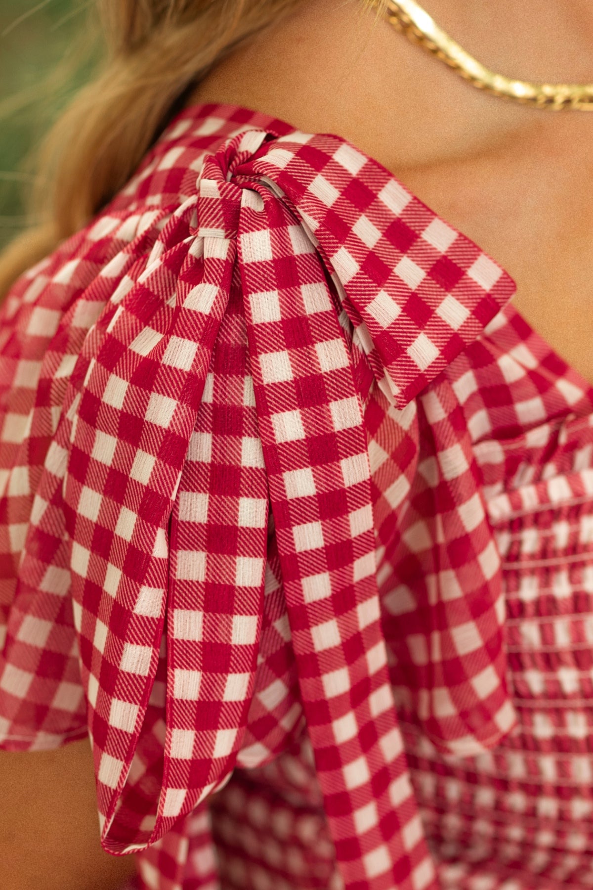 Close-up of a person wearing the Rayna Dress in Red Gingham by Ivy City Co, featuring a smocked bodice, shoulder bow detail, and classic checkered pattern. A gold chain necklace completes the elegant look.