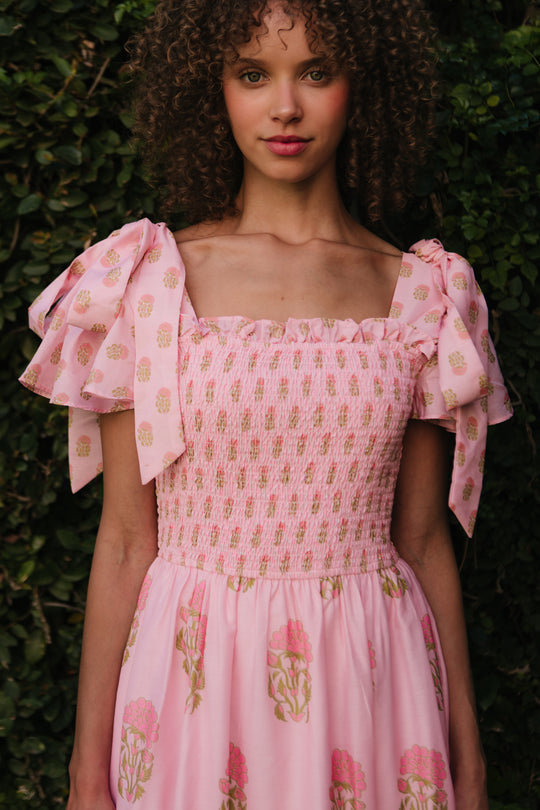 A woman with curly hair stands outdoors before green bushes, wearing Ivy City Cos Rayna Dress in Pink Block Print—featuring floral patterns and puffed sleeves.