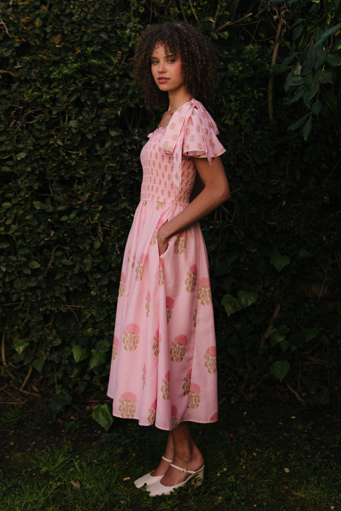 A woman stands outside before leafy greenery in the Ivy City Co Rayna Dress in Pink Block Print—a light pink floral dress with short sleeves, smocked bodice, side pockets, and a maternity-friendly fit. She wears white flats and has curly hair.
