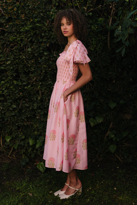 A woman stands outside before leafy greenery in the Ivy City Co Rayna Dress in Pink Block Print—a light pink floral dress with short sleeves, smocked bodice, side pockets, and a maternity-friendly fit. She wears white flats and has curly hair.