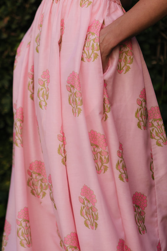 A hand rests in the pocket of Ivy City Co’s Rayna Dress in Pink Block Print, featuring green and pink florals. The blurred greenery background highlights this stylish, maternity-friendly dress option.
