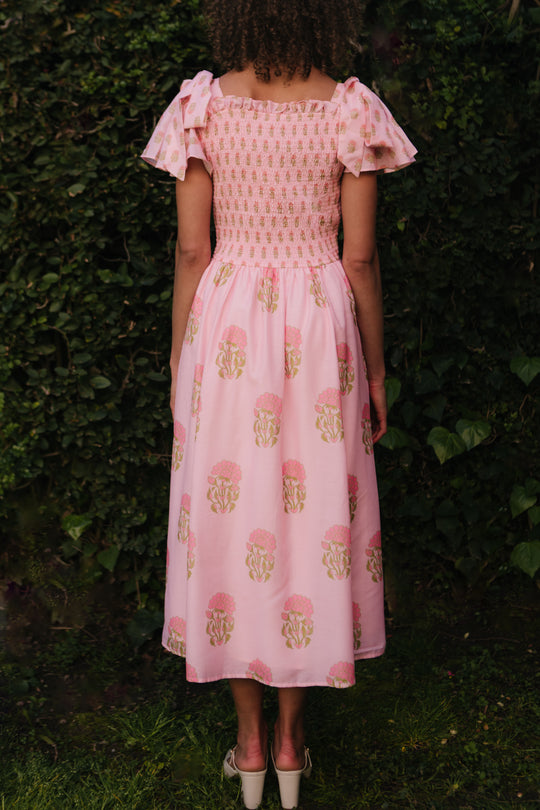 A woman with curly hair faces away outdoors, wearing the Ivy City Co Rayna Dress in Pink Block Print—an ankle-length, maternity-friendly dress with puffed sleeves and floral pattern—set against leafy greenery.