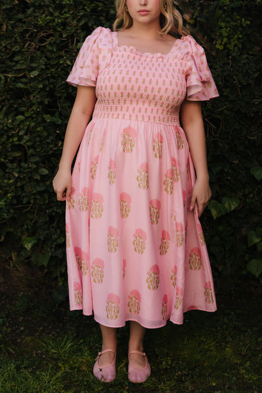 A woman stands outdoors in the Ivy City Co Rayna Dress in Pink Block Print, a maternity-friendly puff-sleeve midi with a smocked bodice, paired with matching pink shoes amidst lush green bushes.