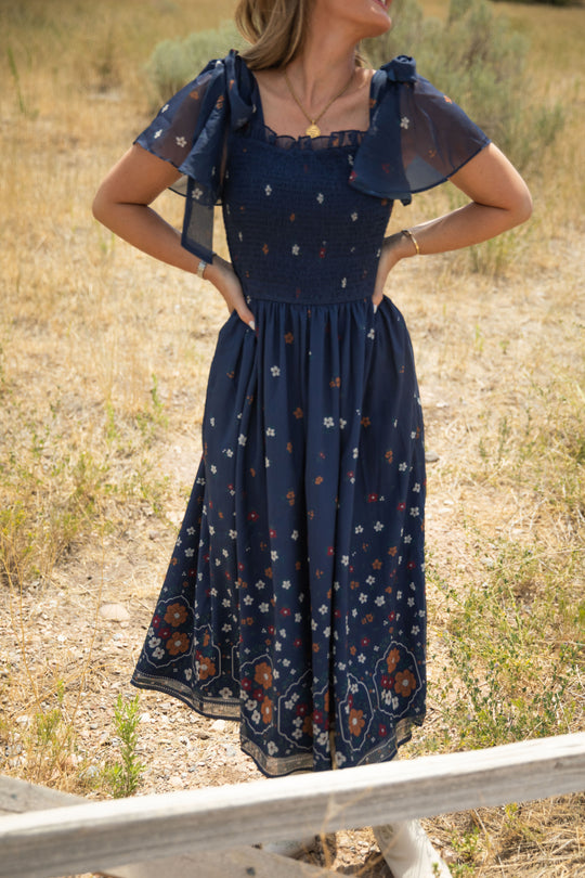 A woman stands outdoors in a field of dry grass and scattered greenery, facing away with her hands on her hips. She is wearing the Ivy City Co Rayna Dress in Midnight Cascading Floral—a blue, short-sleeved chiffon dress featuring a smocked bodice. Her face is not visible.
