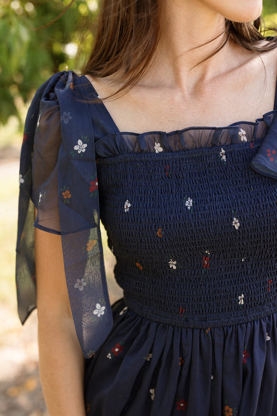A woman in the Rayna Dress in Midnight Cascading Floral by Ivy City Co stands outside, her face turned slightly away. The navy blue, floral chiffon dress features a smocked bodice and sheer puffed sleeves, offering a maternity-friendly fit while maintaining an elegant silhouette.