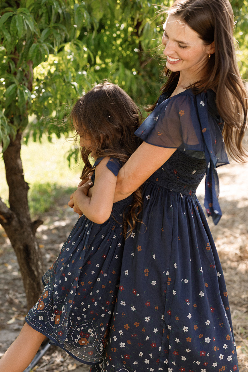 A woman wearing the Ivy City Co Rayna Dress in Midnight Cascading Floral, featuring a smocked bodice and maternity-friendly design, hugs a young girl dressed to match as they smile together outdoors near a tree on a sunny day.