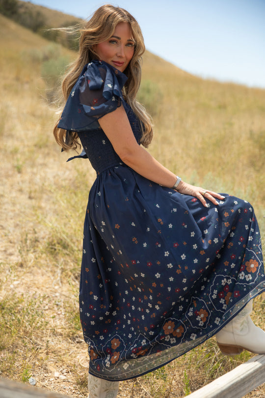 Wearing the Ivy City Co Rayna Dress in Midnight Cascading Floral—a blue chiffon style featuring puff sleeves and a smocked bodice—a woman sits on a wooden fence in a grassy field, gazing over her shoulder. The maternity-friendly silhouette flatters her figure as her long wavy hair falls down her back, complemented by chic white boots.