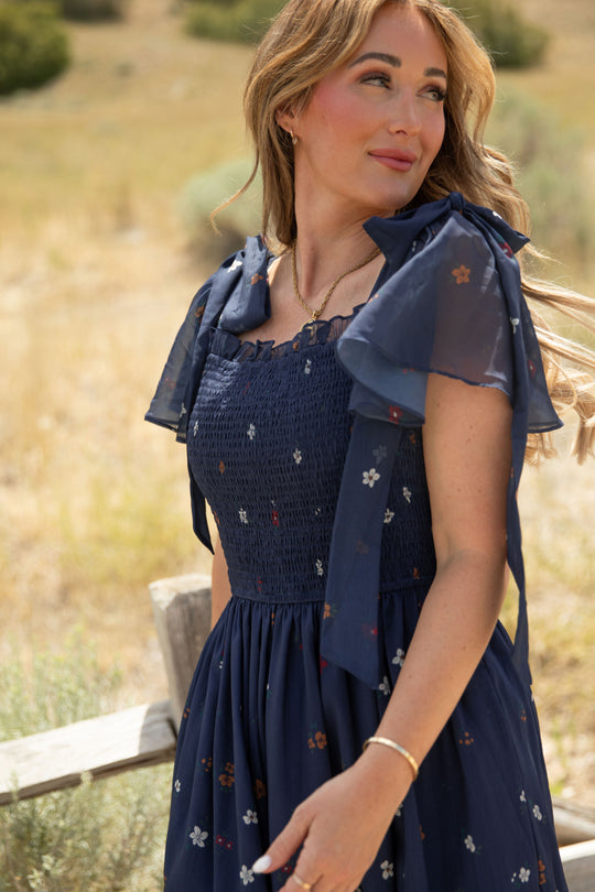 A woman wearing the Ivy City Co Rayna Dress in Midnight Cascading Floral—a navy blue chiffon dress featuring bow sleeves and a smocked bodice—stands outdoors, gazing to the side against a blurred, sunlit field with grass and shrubs.