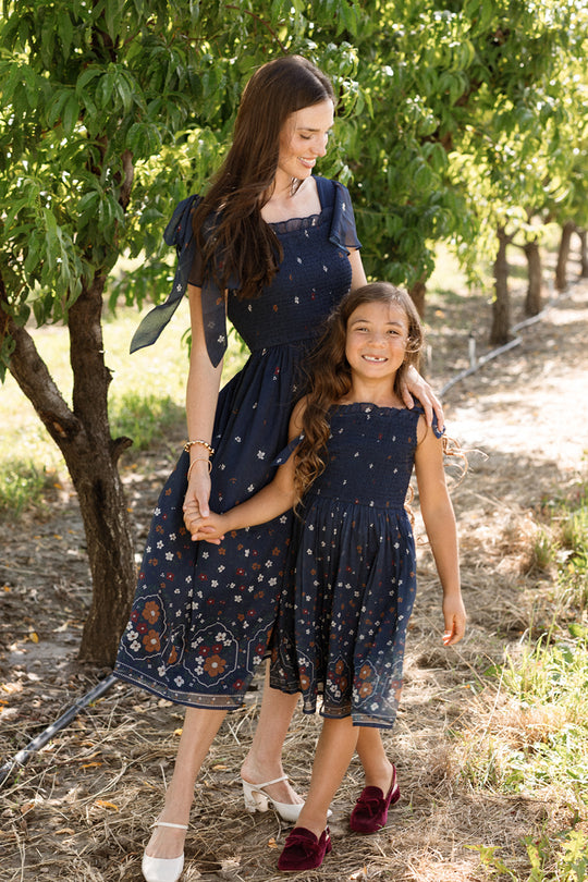A woman and a girl, both wearing the Ivy City Co Rayna Dress in Midnight Cascading Floral—with matching navy blue chiffon fabric, floral patterns, and smocked bodices—hold hands and smile as they stand outdoors among green trees on a sunny day.