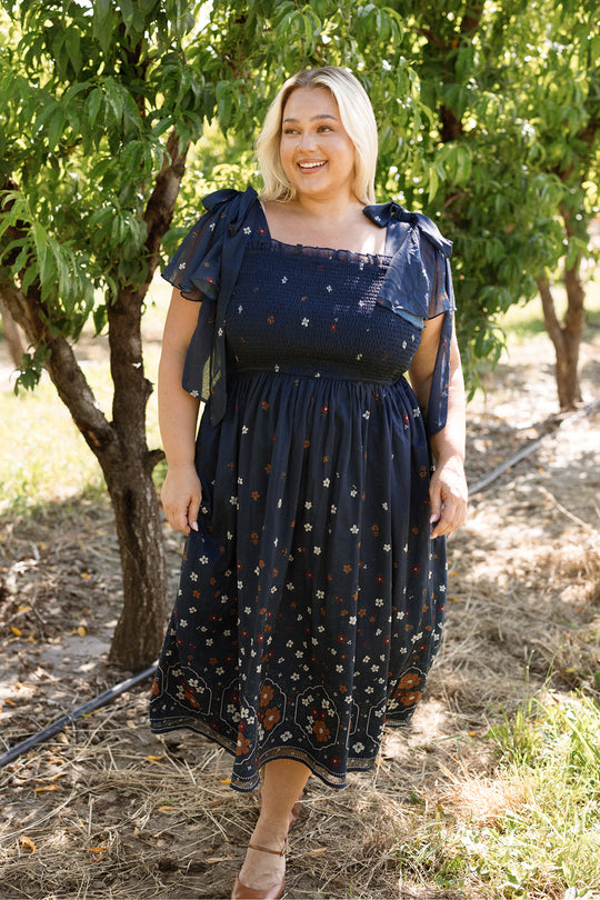 A woman with blonde hair smiles while standing outdoors among green trees on a sunny day, wearing the Ivy City Co Rayna Dress in Midnight Cascading Floral. The dark blue, floral chiffon dress features a smocked bodice and is maternity friendly, offering both comfort and style. She pairs the look with tan flats.