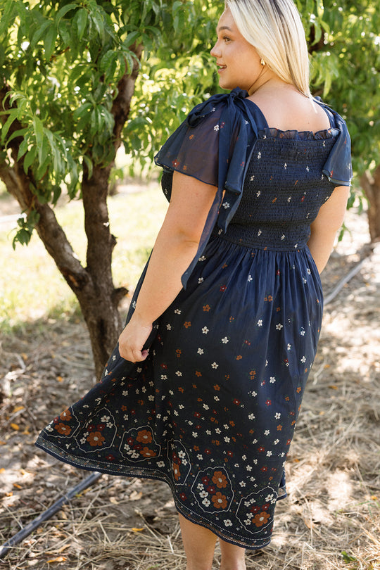Wearing the Ivy City Co Rayna Dress in Midnight Cascading Floral, a woman stands outdoors near trees, softly holding her skirt and looking over her shoulder. Sunlight filters through the leaves, casting a natural, warm glow on the scene, while the smocked bodice of the dress adds an elegant touch to her look.