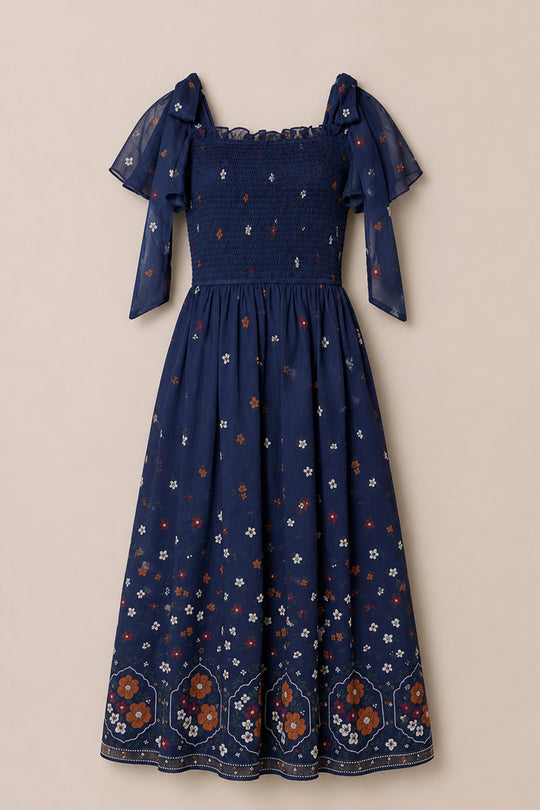 The Rayna Dress in Midnight Cascading Floral by Ivy City Co is a navy blue, short-sleeve chiffon midi dress featuring delicate floral patterns, a smocked bodice, and sheer puff sleeves. Maternity friendly, its hem is detailed with bold red, white, and orange floral designs set against a beige background.