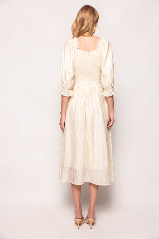 A woman with blonde hair stands facing away, wearing the Ivy City Co Tindra Dress in Cream Sequin—a nursing-friendly midi dress featuring puffed sleeves and ruffled cuffs. She pairs it with nude heeled sandals against a plain, light background.