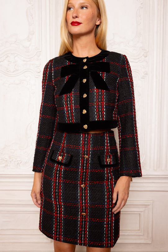 Woman wearing a black and red plaid outfit with a large bow against a white wall.