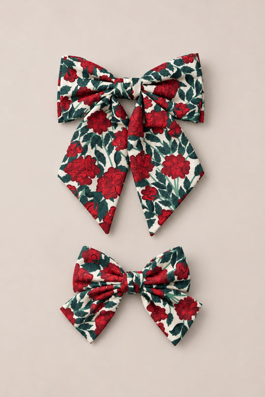 The Ivy City Co “Hair Bows in Red Roses” set features two fabric hair bows adorned with a red rose and green leaf floral print on a cream background. Displayed on a light beige surface, the set includes one larger bow with long tails and one smaller, compact bow—perfect for adding vintage Christmas charm to your look.