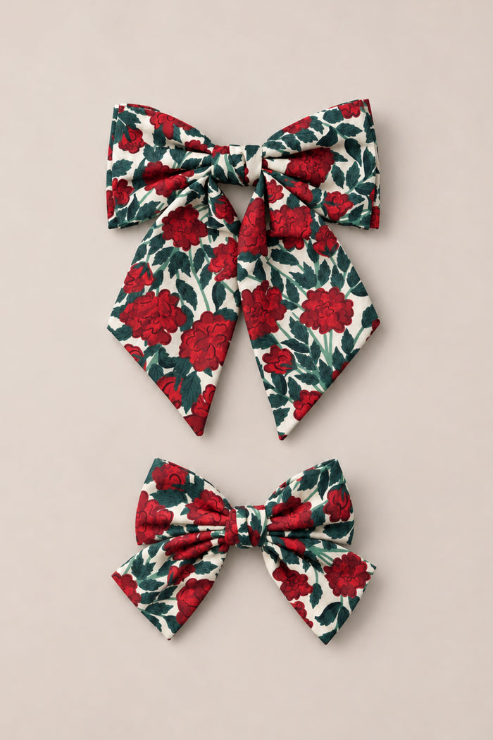 The Ivy City Co “Hair Bows in Red Roses” set features two fabric hair bows adorned with a red rose and green leaf floral print on a cream background. Displayed on a light beige surface, the set includes one larger bow with long tails and one smaller, compact bow—perfect for adding vintage Christmas charm to your look.
