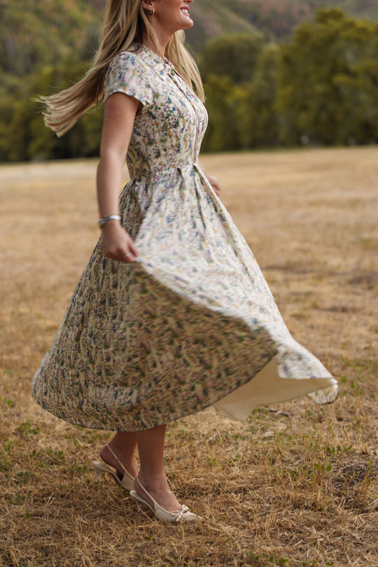 Wearing the Ivy City Co Amber Dress in Autumn Bouquet—a nursing friendly, floral tea-length design—a woman twirls on dry grass in an open field, with softly blurred trees and hills in the background.