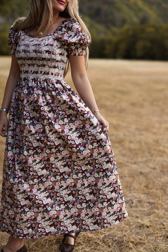 A woman wearing the Ivy City Co Sunday Best Dress in Midnight Garden, featuring puff sleeves and a floral print, stands in a dry grass field holding part of her skirt. Trees and hills are softly blurred in the background. The photo is taken from the shoulders down.