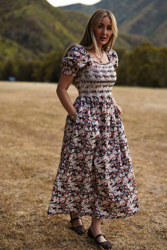 A woman stands outdoors in a dry, grassy field with green hills behind her, wearing the Ivy City Co Sunday Best Dress in Midnight Garden, featuring puff sleeves and a delicate smocked bodice. She pairs it with black shoes and looks toward the camera.