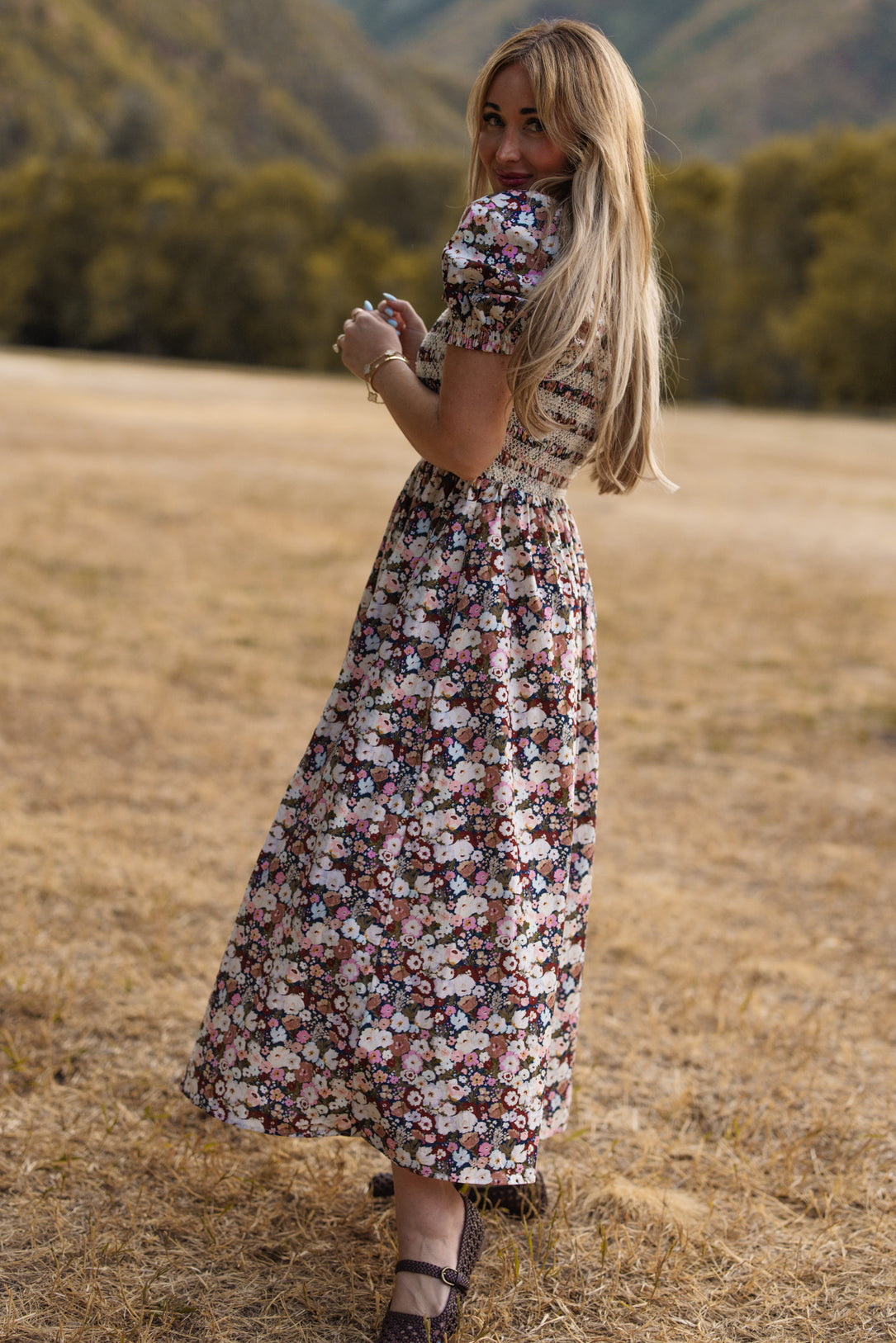 A woman with long blonde hair, wearing the Ivy City Co Sunday Best Dress in Midnight Garden featuring puff sleeves and a smocked bodice, stands on dry grass, smiling over her shoulder with green trees and hills in the background.