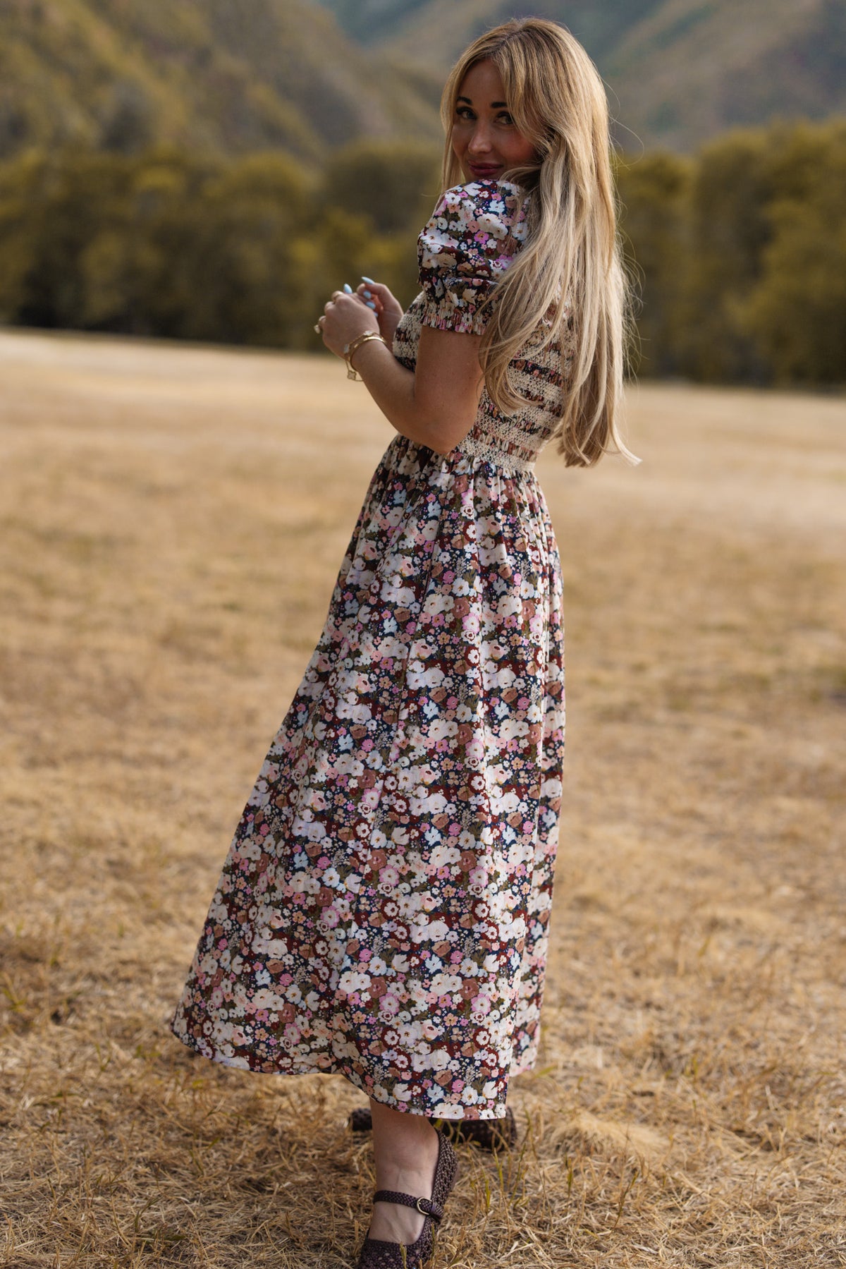 A woman with long blonde hair, wearing the Ivy City Co Sunday Best Dress in Midnight Garden featuring puff sleeves and a smocked bodice, stands on dry grass, smiling over her shoulder with green trees and hills in the background.
