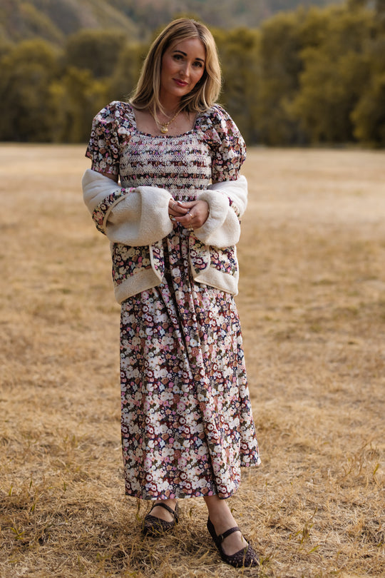 Wearing the Ivy City Co Sunday Best Dress in Midnight Garden—a floral dress featuring a smocked bodice and puff sleeves—paired with a white cardigan, a woman with light brown hair stands on dry grass in a field, surrounded by green trees and hills. She completes the look with black shoes.