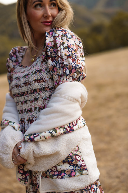 A woman stands outdoors in the Ivy City Co Sunday Best Dress in Midnight Garden, featuring puff sleeves and a smocked bodice, layered under a white sherpa jacket as she gazes off to the side. Blurred grass and trees appear in the background.
