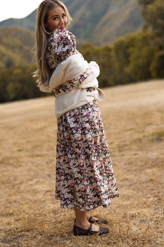 A woman in the Ivy City Co Sunday Best Dress in Midnight Garden, featuring a smocked bodice and floral print, stands on dry grass holding a bundled blanket and smiles over her shoulder. Green trees and hills are visible in the softly blurred background.