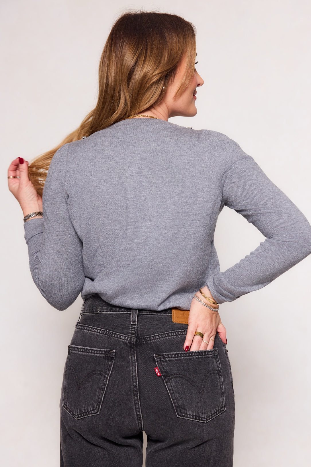 A woman with long light brown hair, wearing the Ivy City Co Rhodes Sweater in Charcoal and high-waisted black jeans, stands facing away from the camera and holds her hair, showcasing effortless fall layering.