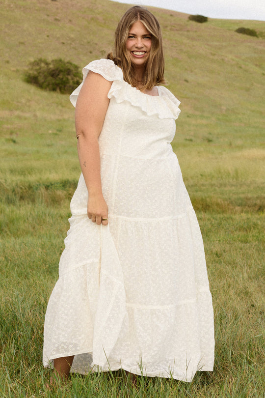 Wearing the Alaina Dress in Ivory by Ivy City Co, a woman stands smiling in a grassy field with hills in the background.