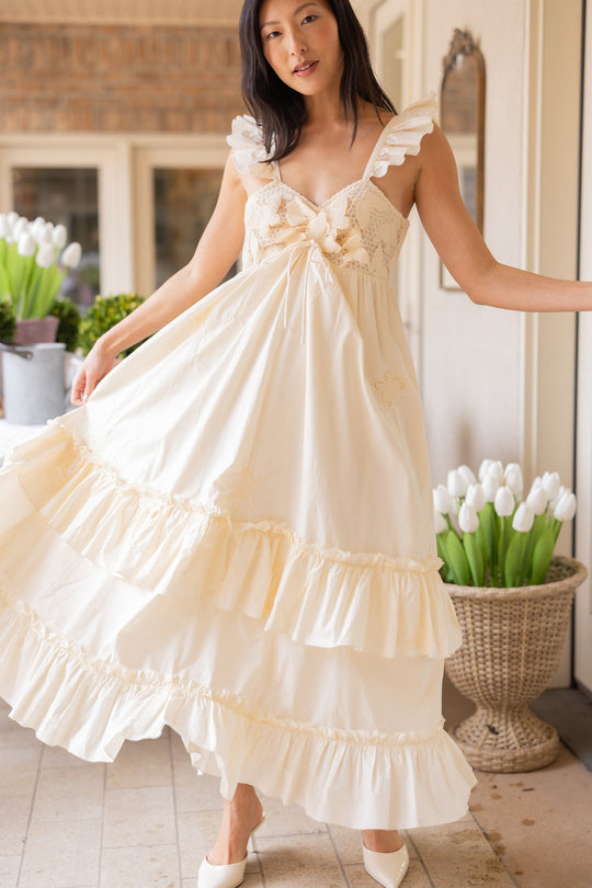 A woman in the Gaia Butterfly Dress in Ivory by Ivy City Co poses indoors near white tulip arrangements, gracefully lifting the skirt of her cream ruffled maxi dress. With long dark hair and standing on a light tiled floor, she creates an elegant look that’s perfect for spring portraits.