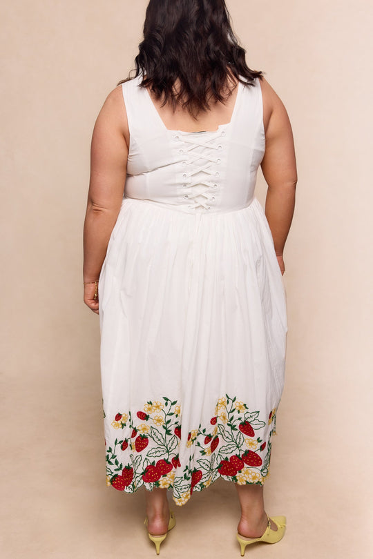 A woman stands facing away, wearing the Ivy City Co Clarissa Dress in Strawberry—a white sleeveless dress featuring a lace-up back and delicate floral embroidery at the hem. She has wavy dark hair and is paired with yellow shoes.