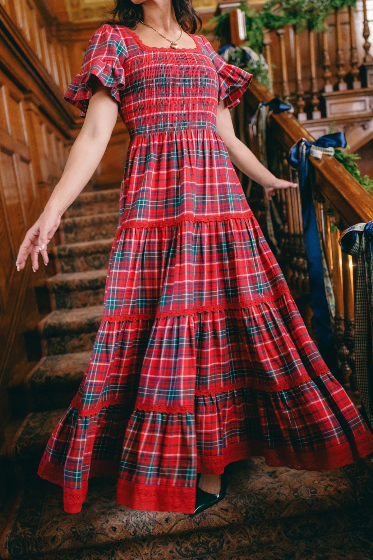 Wearing the Ivy City Co Rosario Dress in Christmas Cheer Plaid, a person stands on a carpeted staircase in a wood-paneled interior with their arms slightly outstretched—a festive and charming choice for a family photo outfit.
