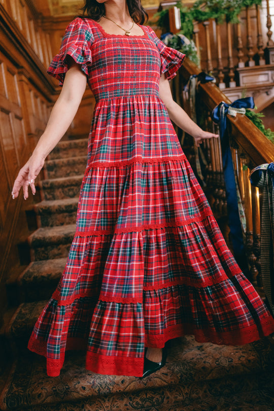 Wearing the Ivy City Co Rosario Dress in Christmas Cheer Plaid, a person stands on a carpeted staircase in a wood-paneled interior with their arms slightly outstretched—a festive and charming choice for a family photo outfit.