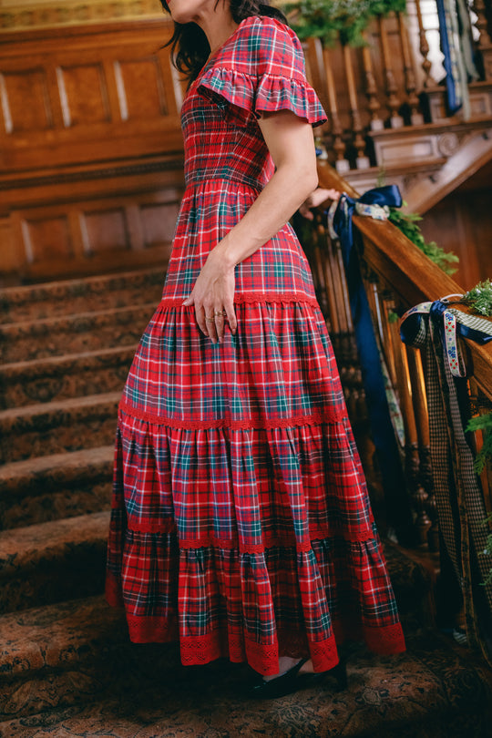 A woman wearing the Ivy City Co Rosario Dress in Christmas Cheer Plaid stands on a carpeted staircase, holding the wooden railing. Her face is partially out of frame, and the background features ornate wood paneling—making this an ideal family photo outfit.