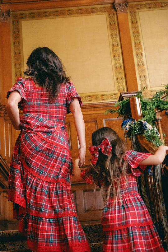 A woman and a girl, both dressed in matching Ivy City Co Rosario Dresses in Christmas Cheer Plaid, walk up a wooden staircase adorned with festive greenery and ribbons—a perfect family photo outfit.