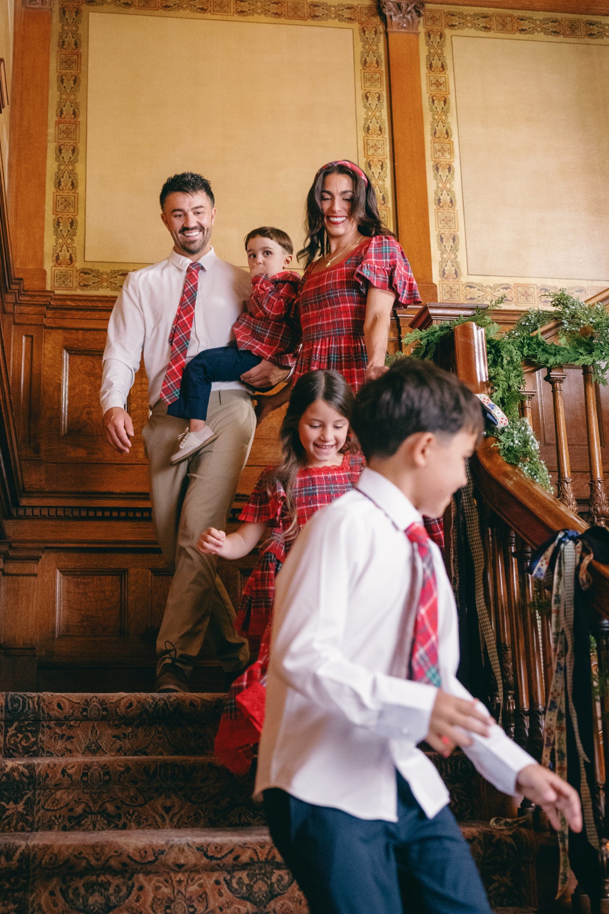 A smiling family in coordinated red and white outfits, including the Ivy City Co Rosario Dress in Christmas Cheer Plaid—perfect for family holiday photos—walks down a carpeted staircase adorned with garland, with wood paneling and ornate walls in the background.