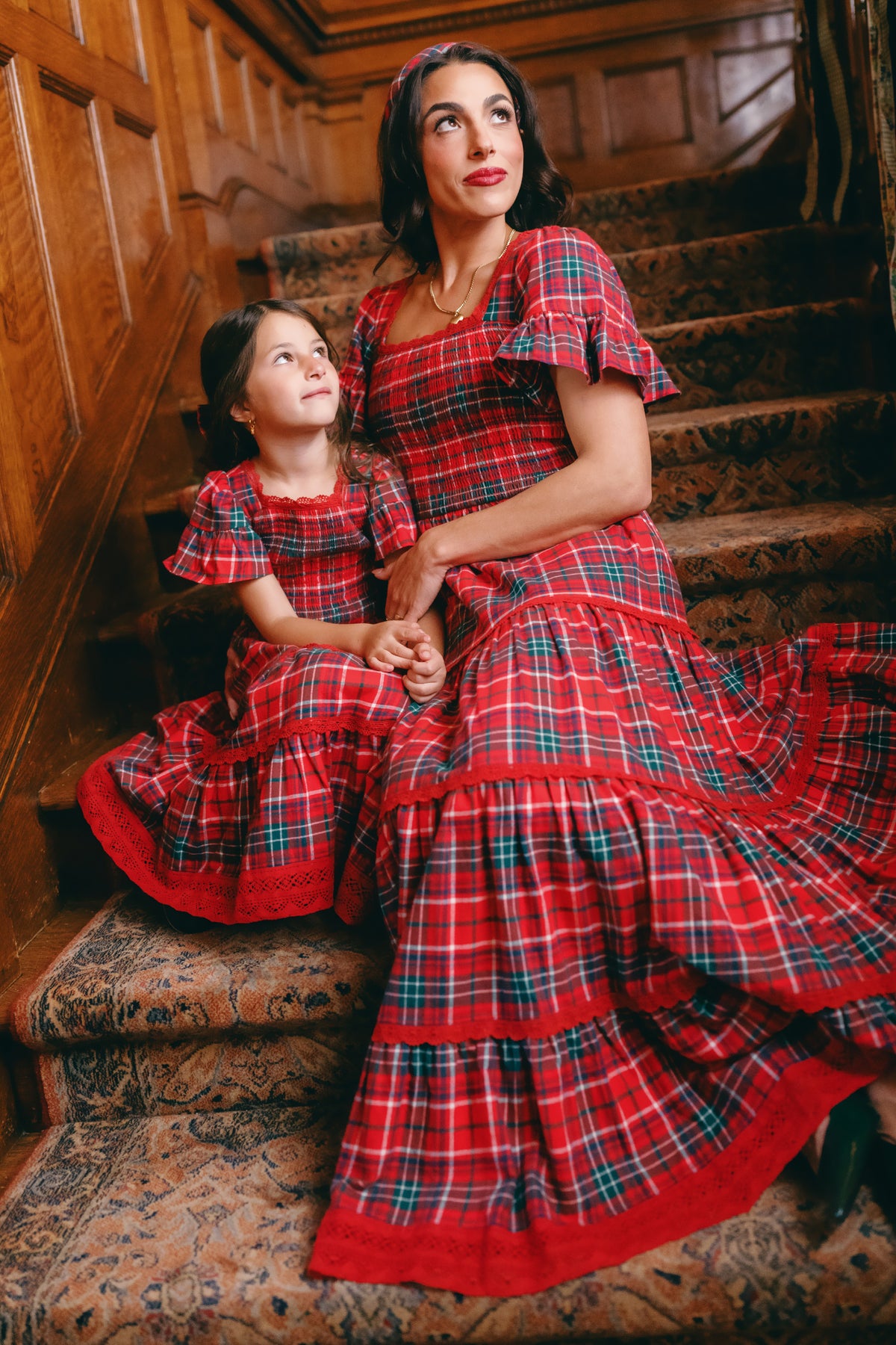 A woman and a young girl sit on carpeted stairs, both dressed in matching Ivy City Co Rosario Dresses in Christmas Cheer Plaid. The woman looks ahead while the girl gazes up at her, both smiling softly. Wood paneling lines the wall behind them—making these coordinated outfits the perfect choice for a festive family photo.