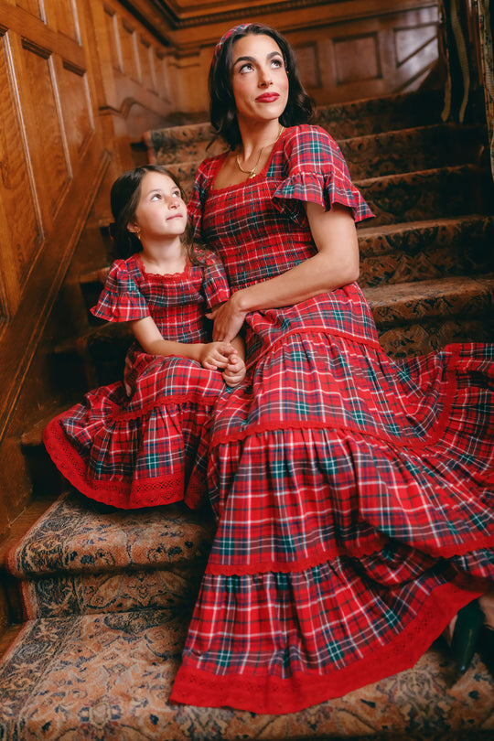 A woman and a young girl sit on carpeted stairs, both dressed in matching Ivy City Co Rosario Dresses in Christmas Cheer Plaid. The woman looks ahead while the girl gazes up at her, both smiling softly. Wood paneling lines the wall behind them—making these coordinated outfits the perfect choice for a festive family photo.